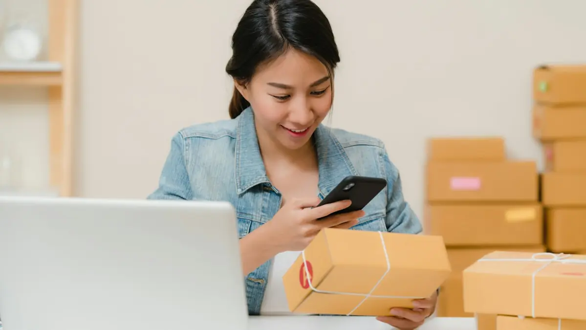 Vendedora conferindo um pedido no celular, com caixas prontas para envio